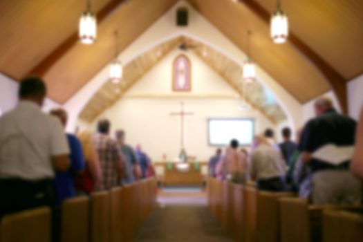 Blurred Photo of the Iterior of a Church Sanctuary with Congregation, Pastor, and a Large Cross Visible A blurred photo of the inside of a church sanctuary that is filled with people in the pews, and the pastor stands under a large cross at the altar.