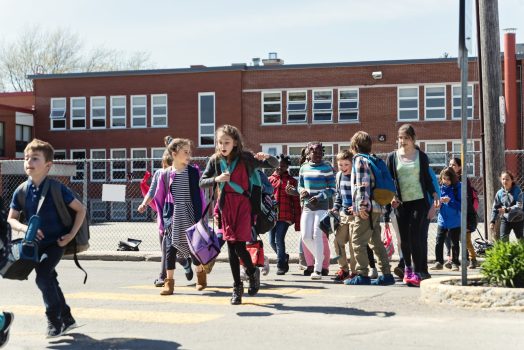 Large group of multi-ethnic elementary age school kids crossing the street getting out of school. Most of them are carrying backpack and lunch box. Some are running. Horizontal full length outdoors shot on a bright sunny day with copy space.