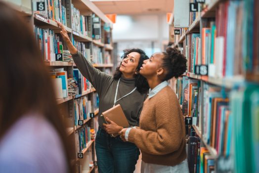 Hispanic student searching a specific book in a library. Walking in between the book shelves and smiling.