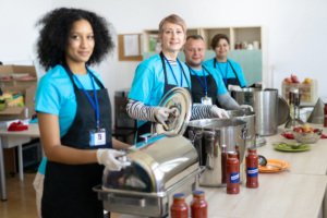 Multi-racial volunteers standing in a line at a table ready to serve homeless people with a hot meal, wearing matching t-shirts, protective aprons and gloves for hygiene