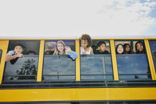 Medium wide shot of smiling middle school kids looking out open windows of school bus