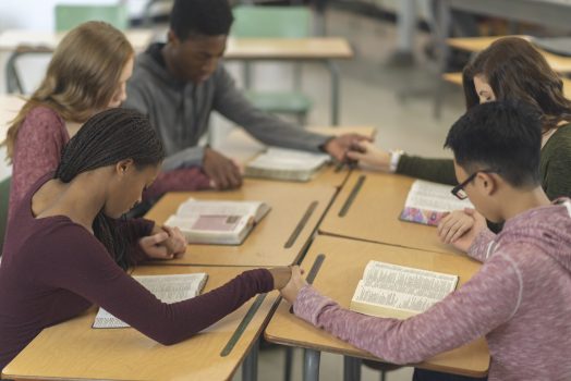 Five students in a classroom are sitting with their desks pushed together. They are holding hands in a circle and have their heads bowed in prayer. There are bibles open on their desks.