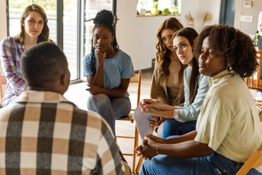 Selective focus shot of diverse group of young people sitting in a circle and listening, with compassion, to a young man sharing his problems during a group therapy session.