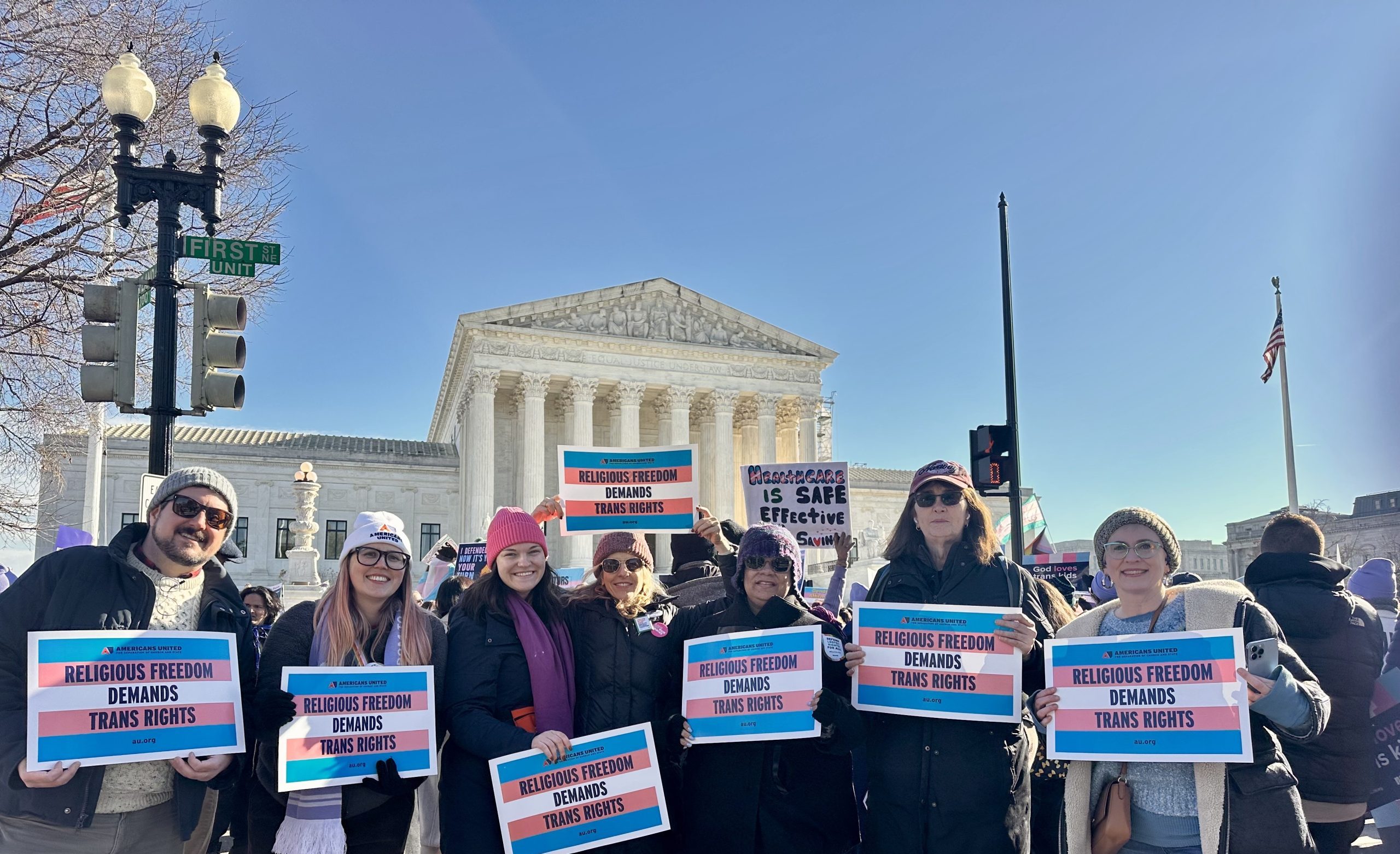 a group of diverse people standing together in front of the U.S. Supreme Court holding rally signs supporting trans rights.